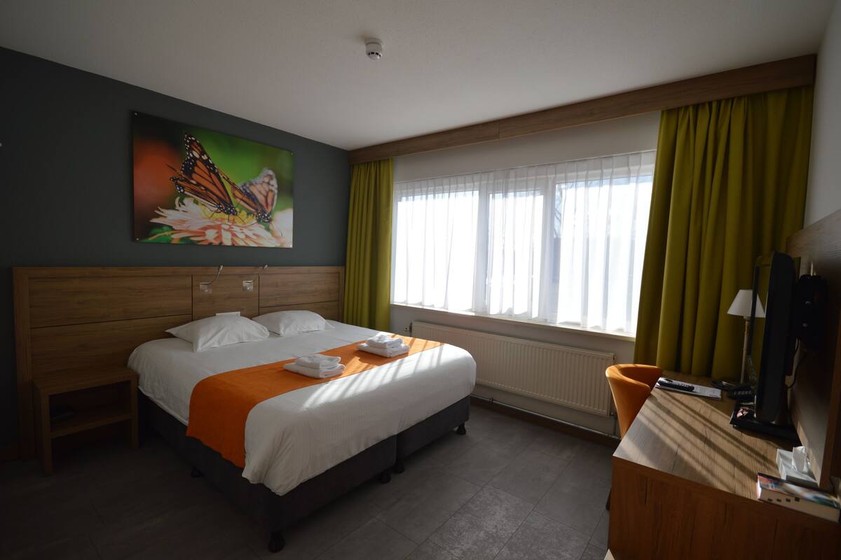 A hotel room with a bed, a desk, a chair, a painting, and a window with curtains.