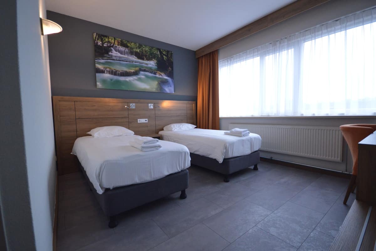 A hotel room with two beds, a painting of a waterfall, a window with curtains, and a radiator.