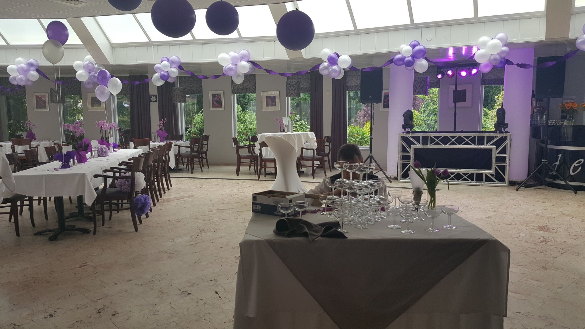 A banquet hall with tables set for dining, a stage with speakers, and a ceiling with hanging balloons.
