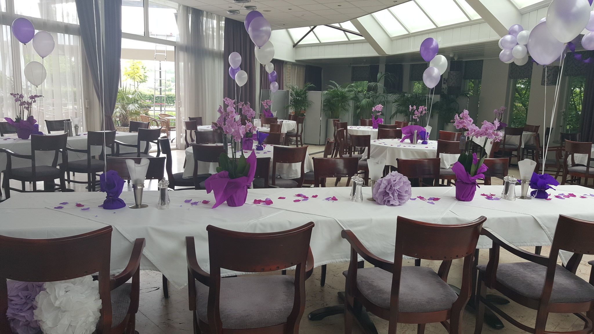 Een balzaal met ronde tafels, bedekt met witte tafelkleden, paarse en witte ballonnen en bloemstukken als decoratie.