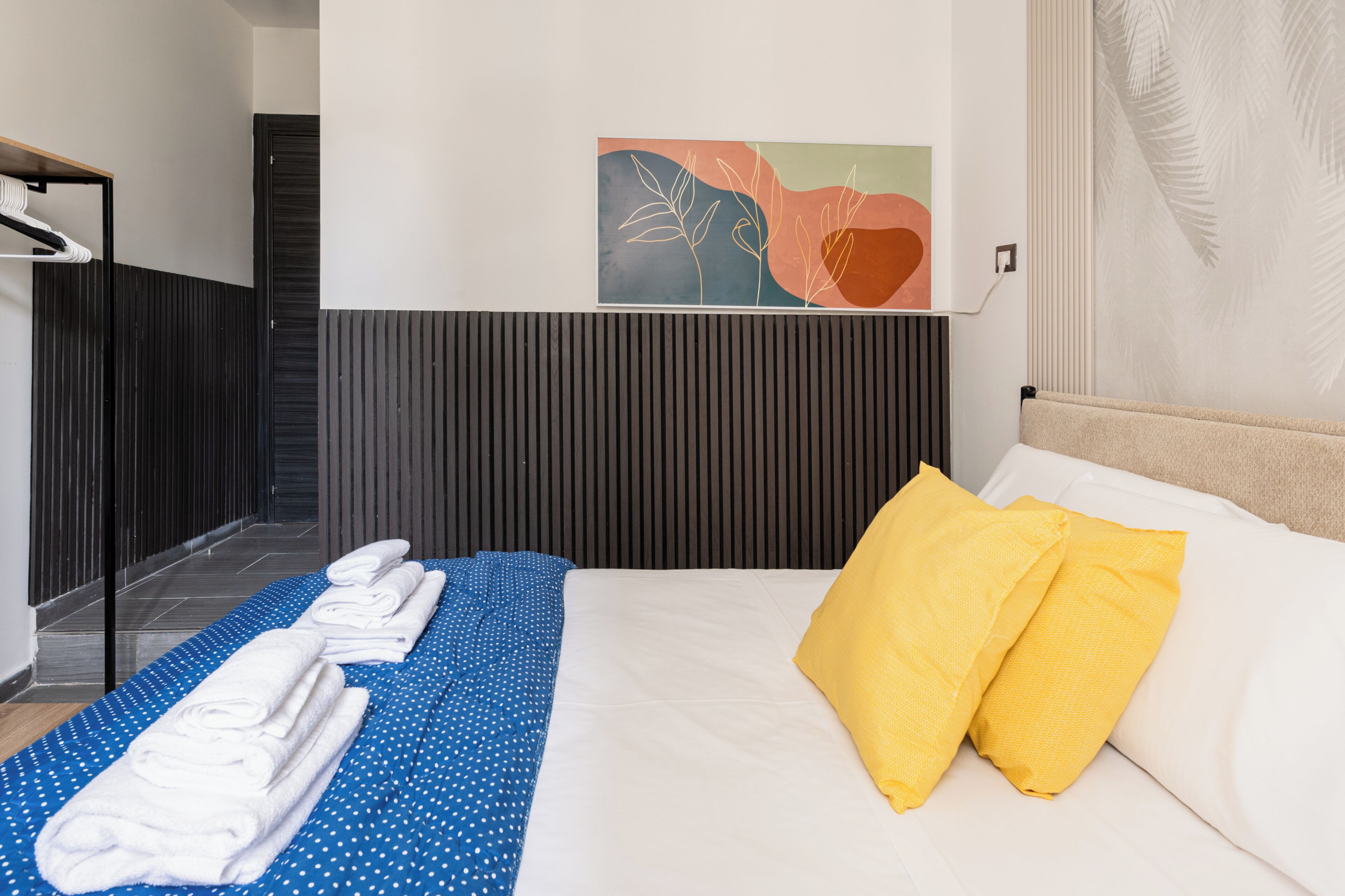 Une chambre d’hôtel avec un lit, une couvre-lit à pois bleus, des serviettes blanches, un oreiller jaune, une œuvre d’art murale et un mur recouvert de panneaux en bois.