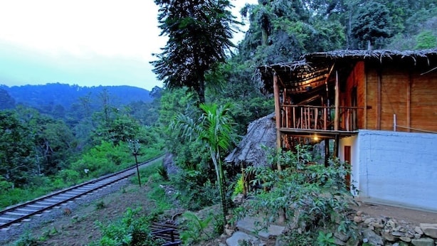A rustic wooden cabin with a thatched roof, surrounded by lush greenery and a railway track. - Ella Bamboo Cabana (Ella)