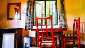 A room with a wooden table and chairs, a window with a metal screen, a small trash bin, and a refrigerator. - Ella Bamboo Cabana (Ella)