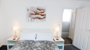 A bedroom with a bed, two bedside tables with lamps, a white door, and a hallway. - Church View Apartments (Weymouth)