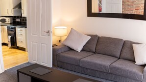 A modern living room with a grey sofa, a small table, and a kitchen visible through an open door. - Church View Apartments (Weymouth)