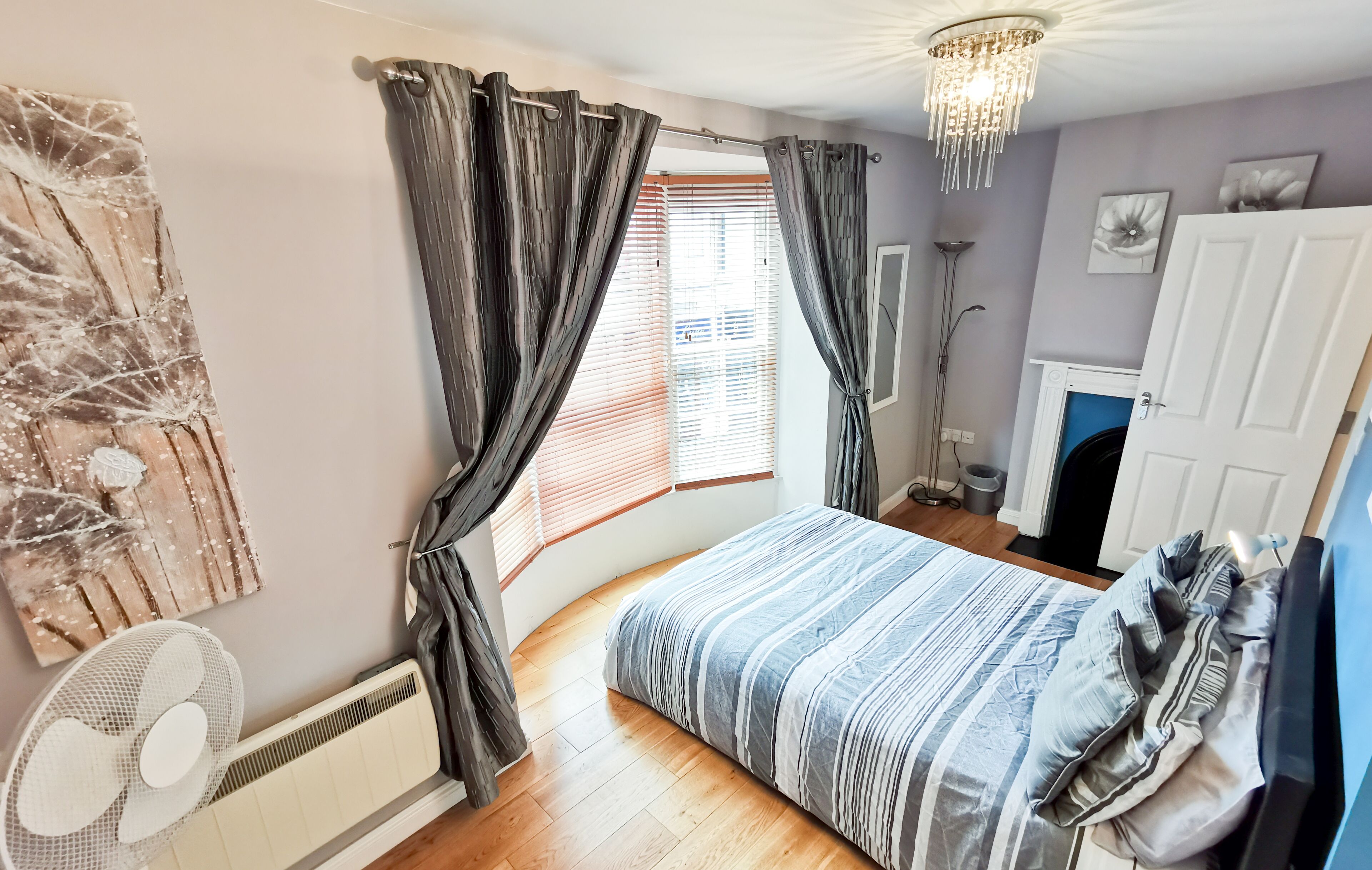 A bedroom with a bed, curtains, a radiator, a fan, a fireplace, and a door.