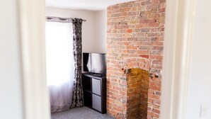 A room with a brick fireplace, a TV on a black cabinet, and a window with a curtain. - Church View Apartments (Weymouth)