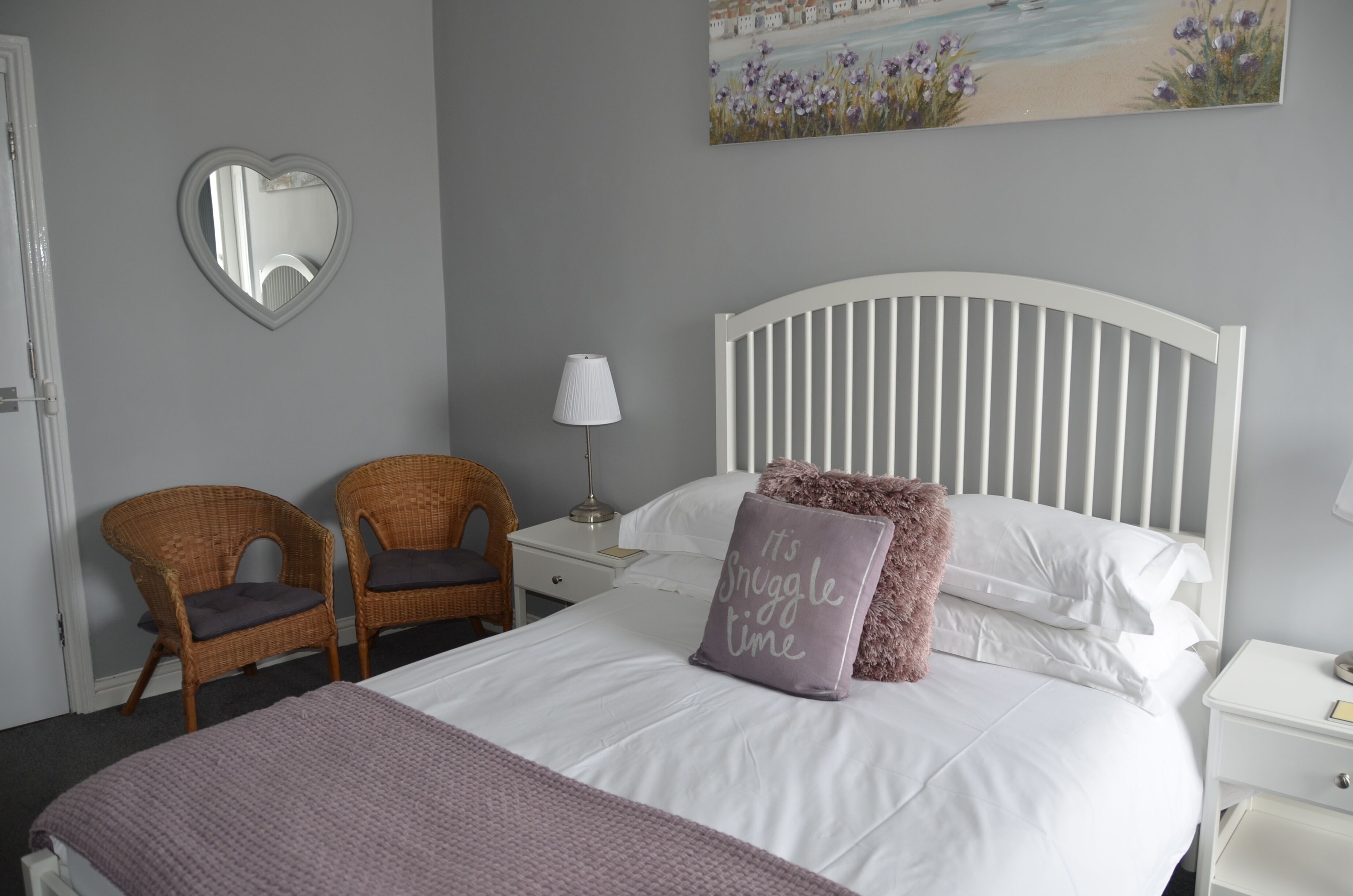 A neatly arranged bedroom with a white bed, two wicker chairs, a nightstand with a lamp, and a heart-shaped mirror on the wall.