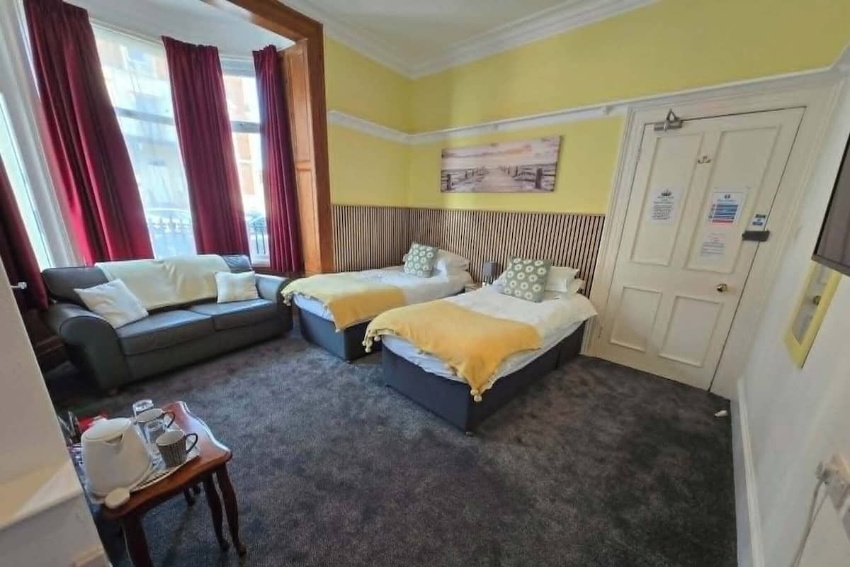 A hotel room with two beds, a sofa, a small table with a cup and saucer, and a window with curtains.
