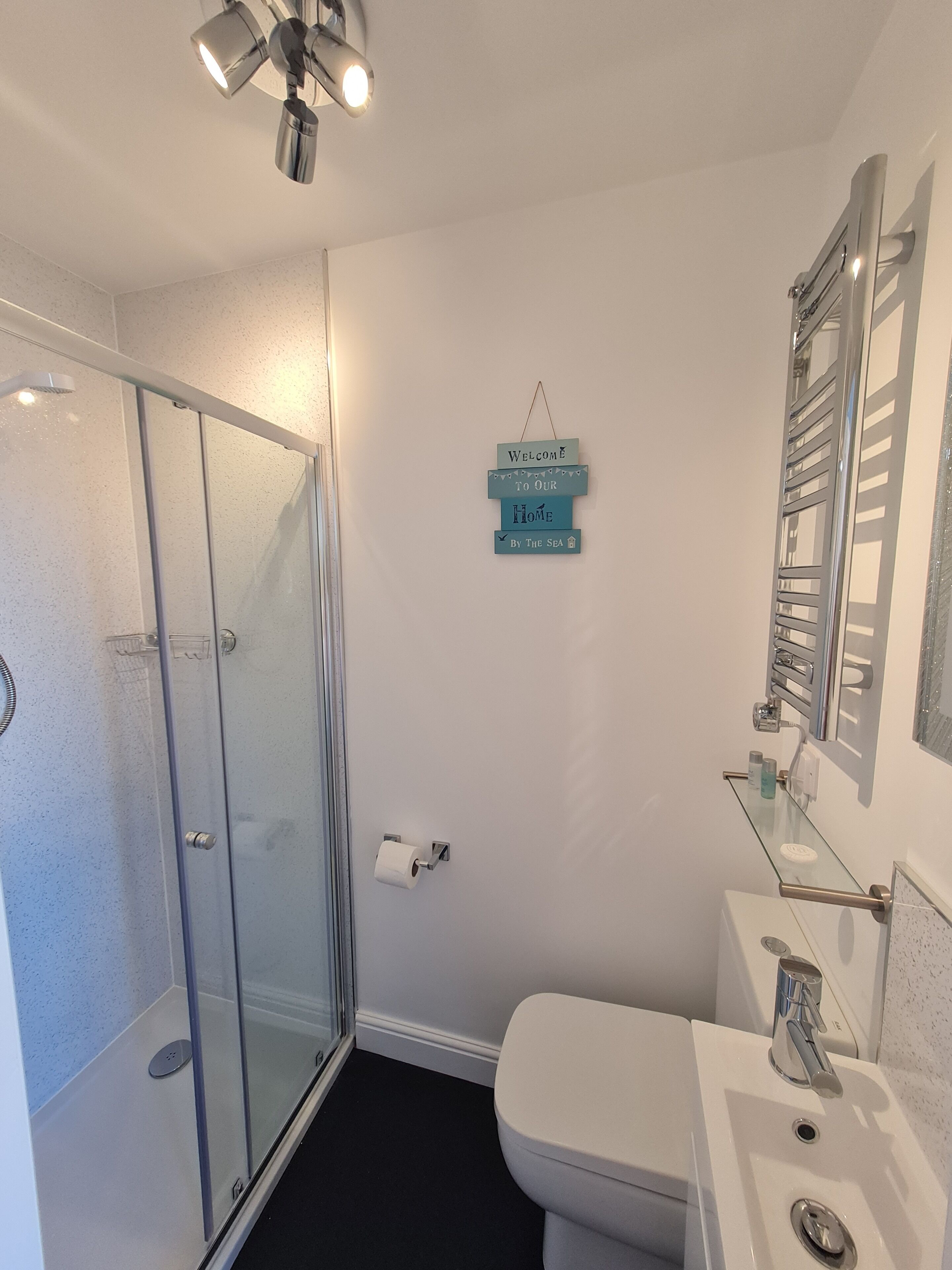 A bathroom with a glass shower enclosure, a toilet, a sink with a mirror, and a welcome sign on the wall.