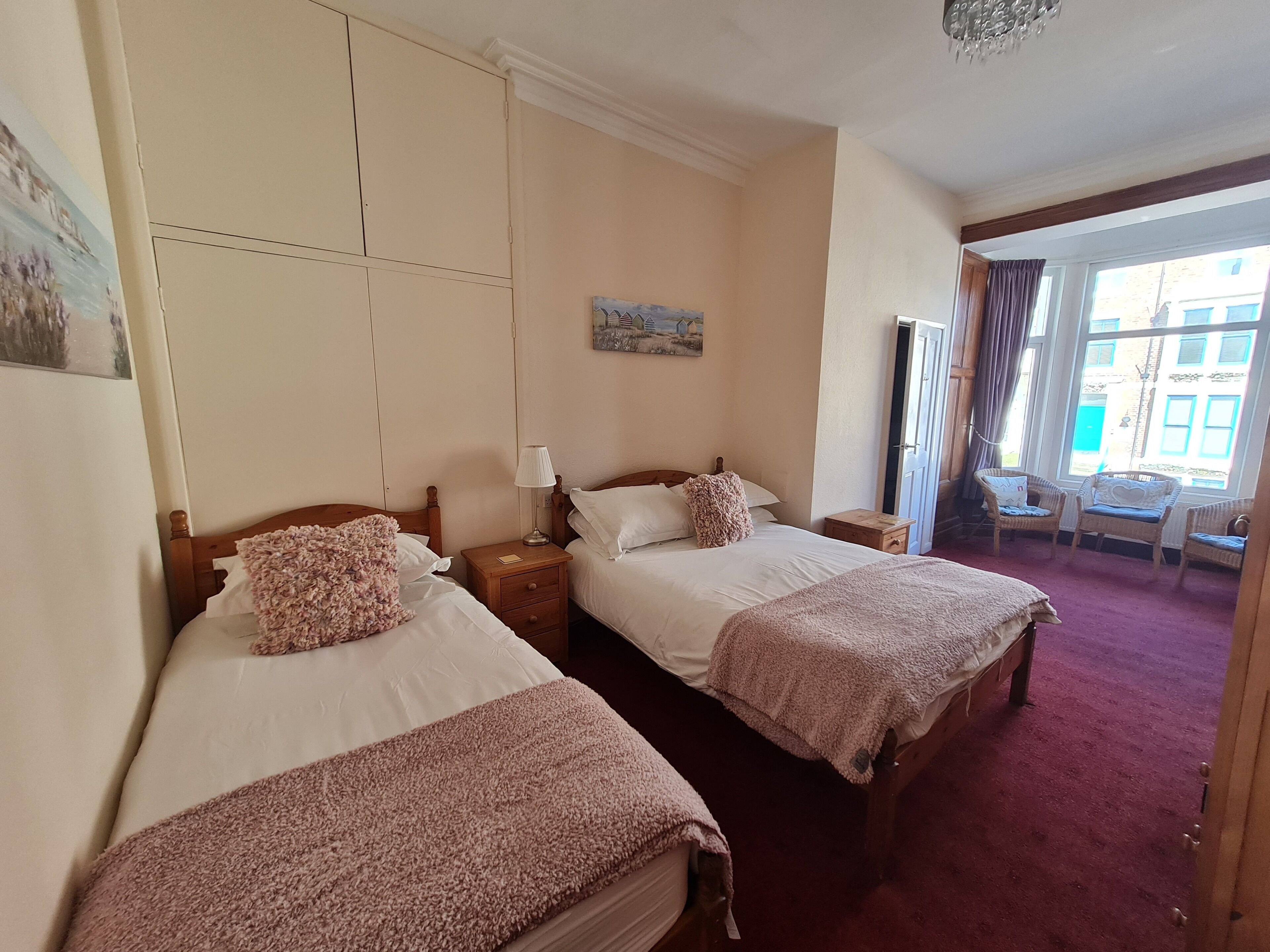 A hotel room with two beds, a small table, a lamp, and a view of the outside through a window.