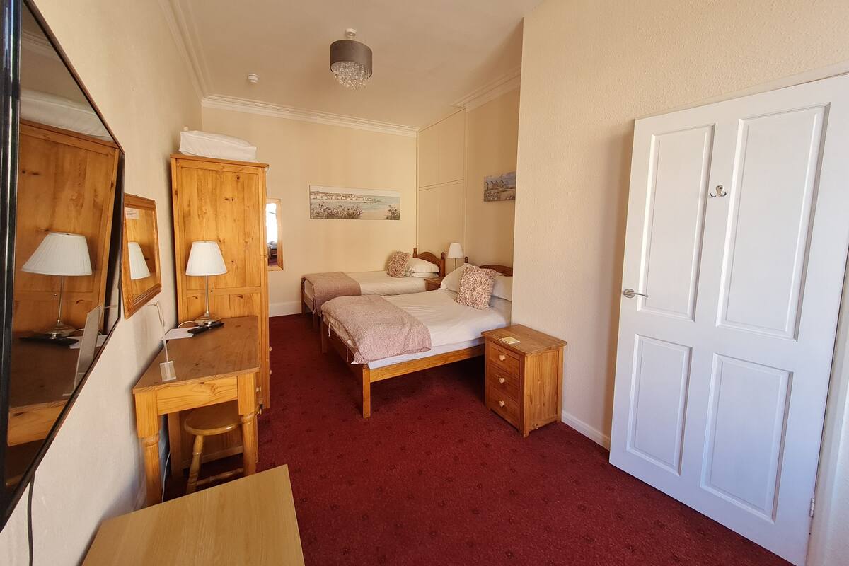 A bedroom with a wooden desk, two beds, a wardrobe, and a mirror.