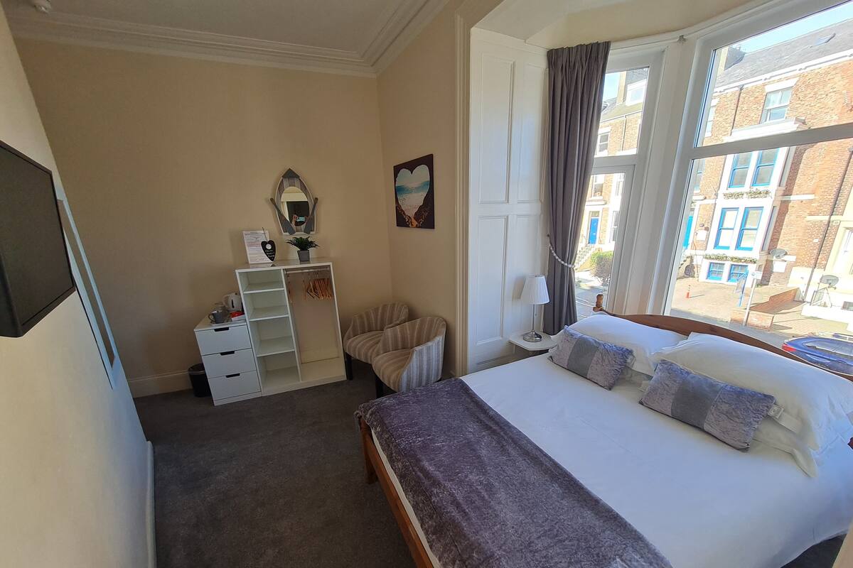 A hotel room with a bed, a desk, a chair, a wardrobe, and a window with a view of buildings.