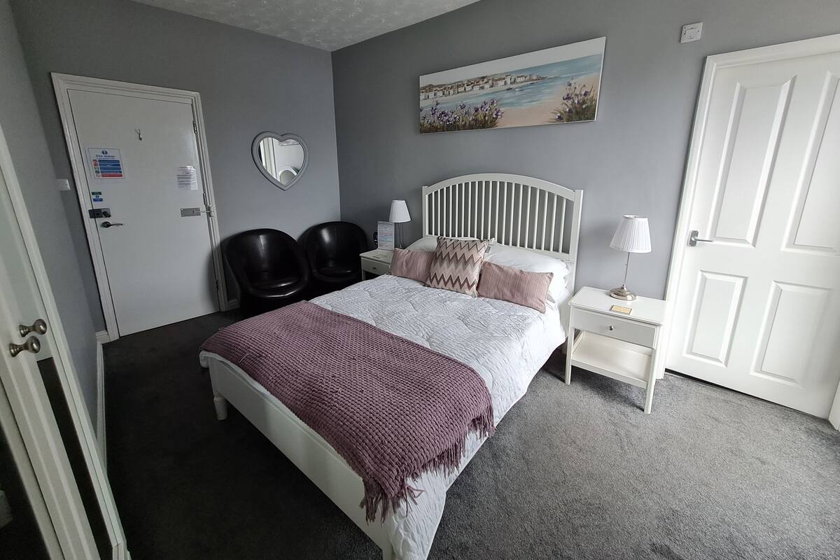 A bedroom with a bed, two chairs, a nightstand, a painting, and a door.