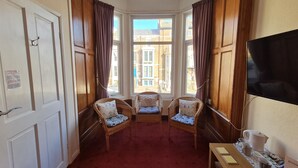Una habitación de hotel con alfombra roja, dos sillones de mimbre con cojines, una mesa de madera con una jarrón y vasos, y un televisor en la pared.