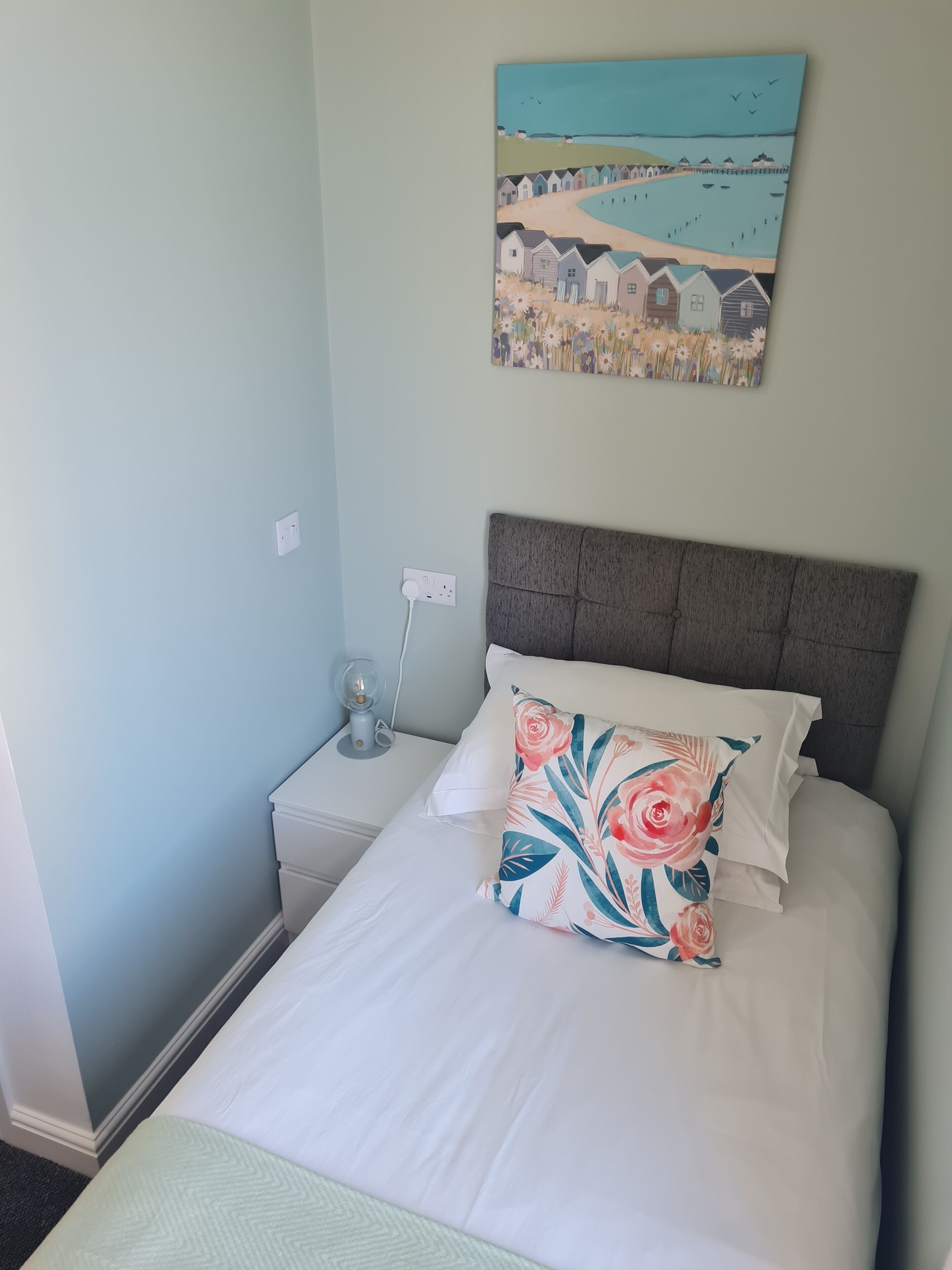A bedroom with a bed, a nightstand, a wall-mounted painting, and a power outlet.