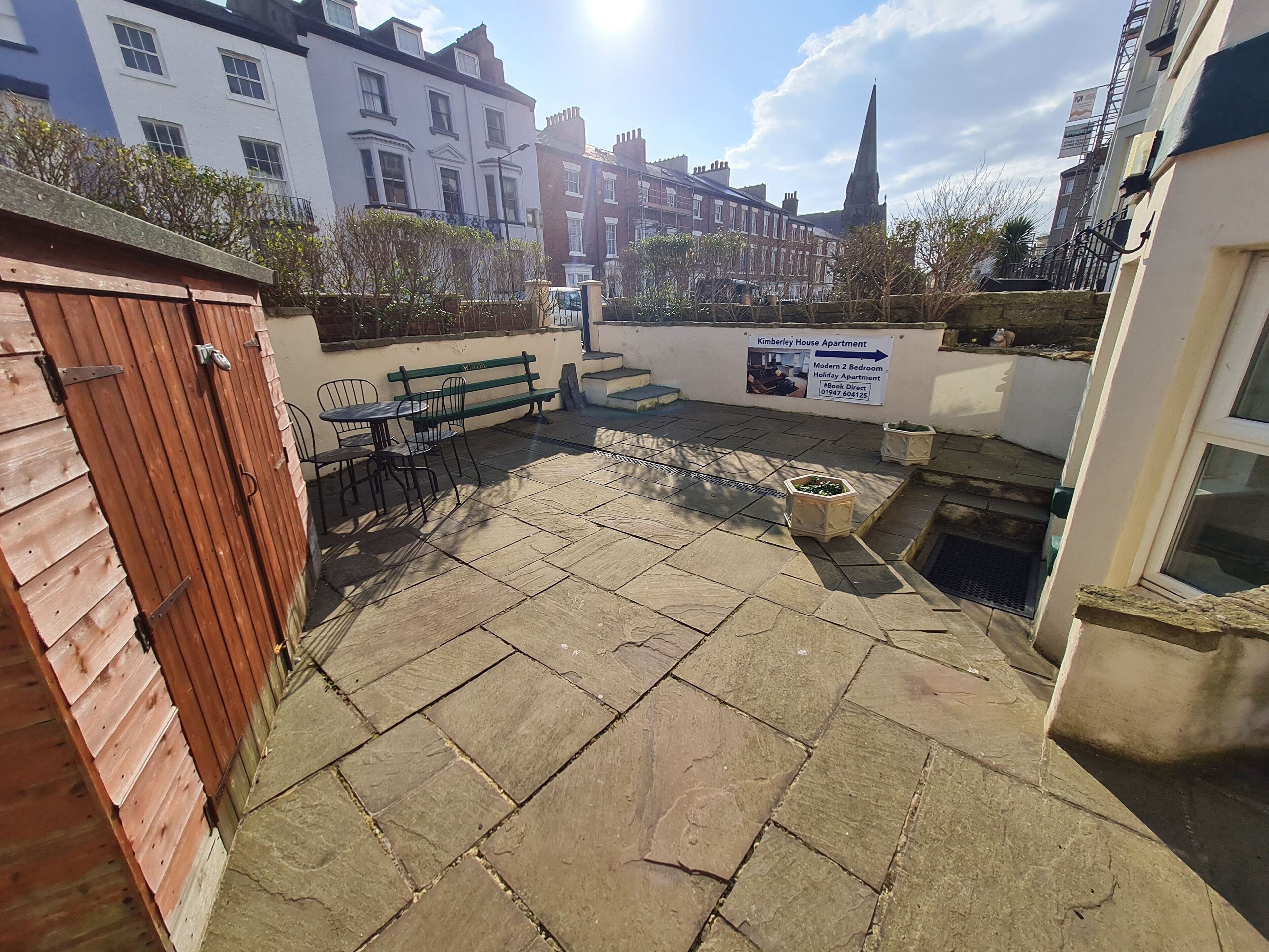 A courtyard with stone flooring, a wooden gate, and a bench. There is a sign indicating a modern 2 bedroom holiday apartment.