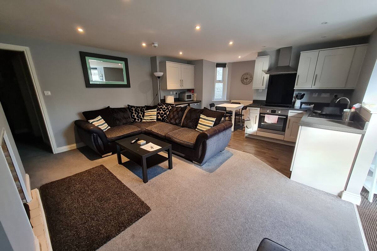 A modern living room with a sectional sofa, a coffee table, and a kitchen area with white cabinets and a stove.