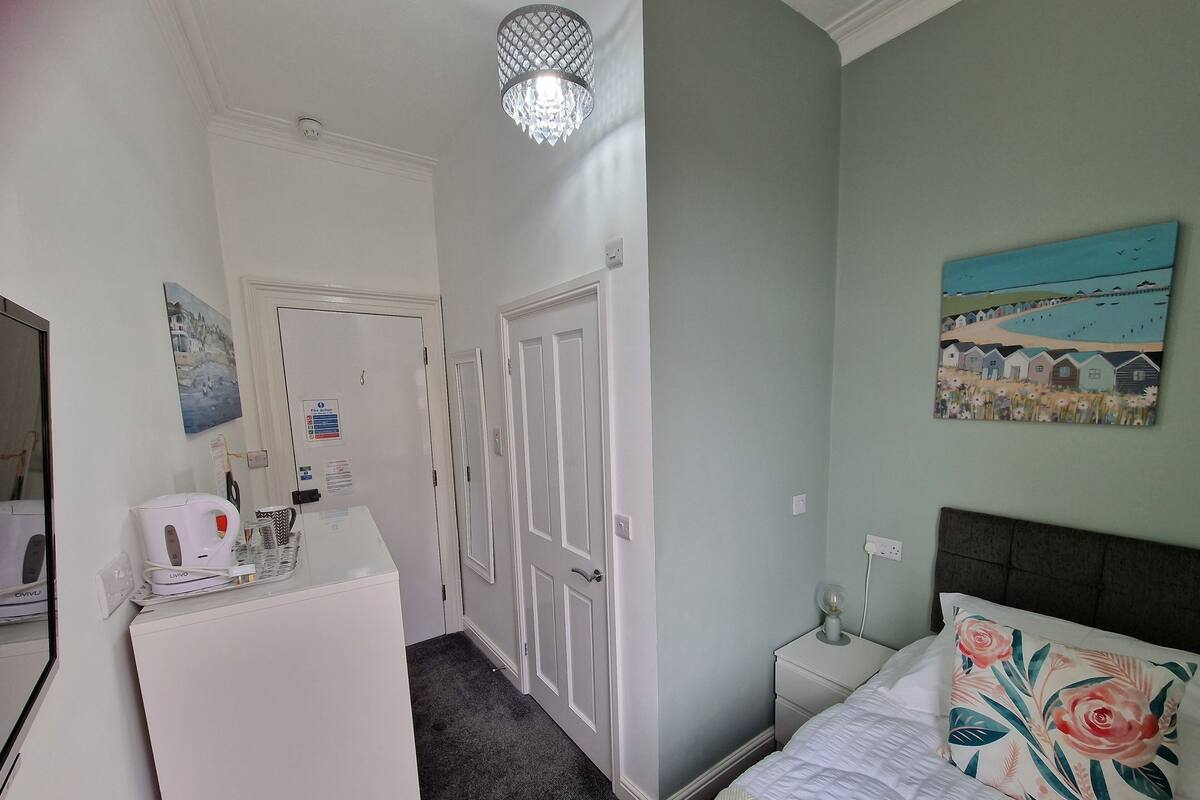 A bedroom with a bed, a nightstand, a lamp, a painting, a door, and a kitchenette area with a kettle and a coffee maker.