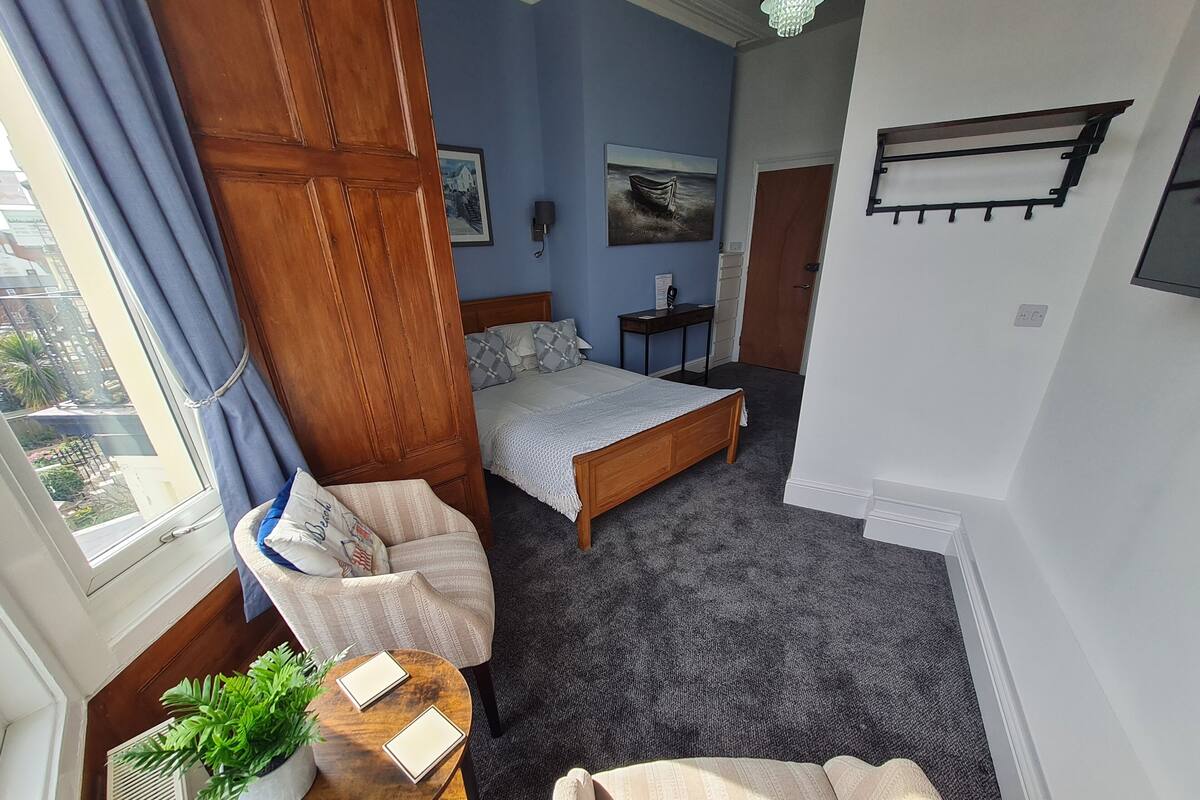 A bedroom with a bed, a chair, a small table with a plant, and a view of the outside through a window.