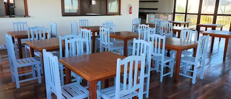 A dining area with multiple wooden tables and light blue chairs, large windows, and a hanging light fixture.