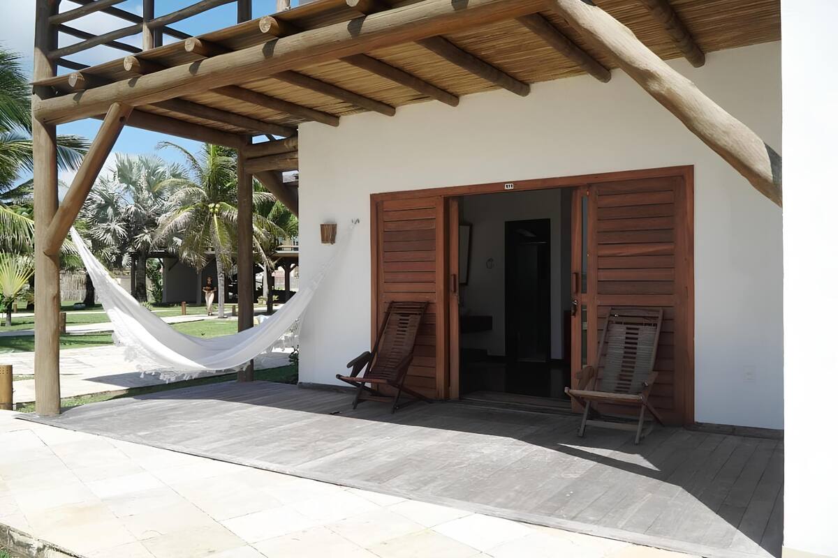 Une pergola en bois avec un hamac, deux chaises en bois et une entrée d’immeuble dotée de volets en bois.