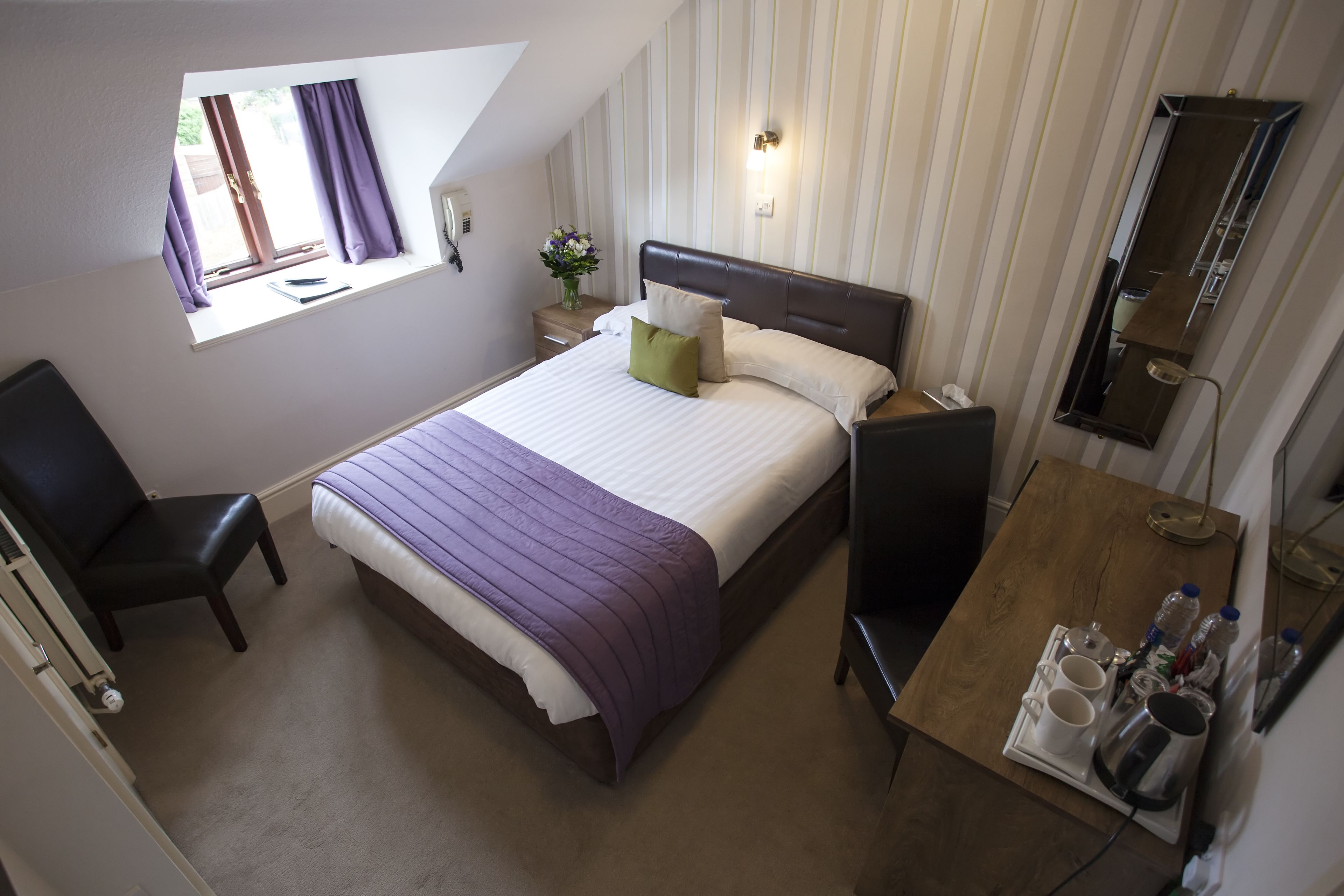 A bedroom with a bed, a chair, a bedside table with a lamp, and a small table with cups and bottles.