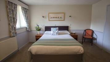 A hotel room with a large bed, a nightstand, a chair, a window with curtains, a radiator, and a framed picture on the wall.