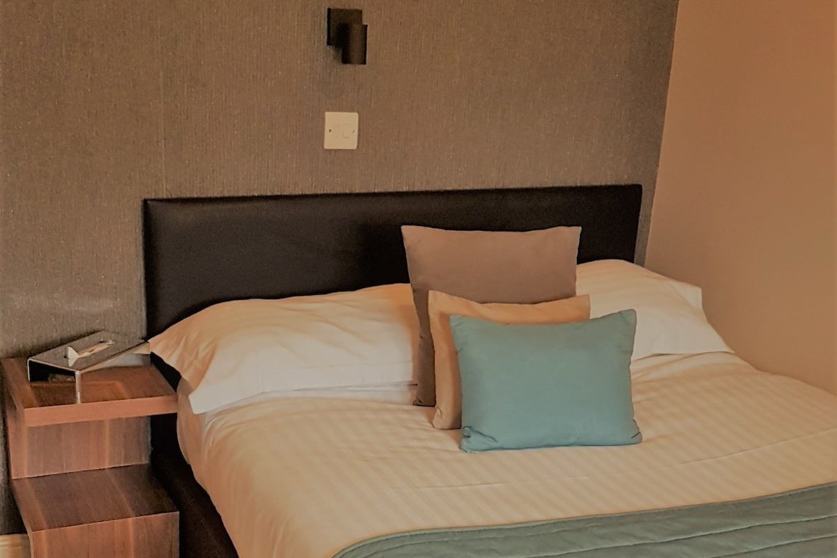 Ein ordentlich bezogenes Bett mit einer hellblauen Decke, ein Nachttisch mit einer Lampe und ein an der Wand befestigter Lichtschalter.