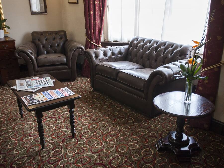 A living room with a brown leather sofa, a wooden coffee table with newspapers, a patterned carpet, and a small round table with a vase.