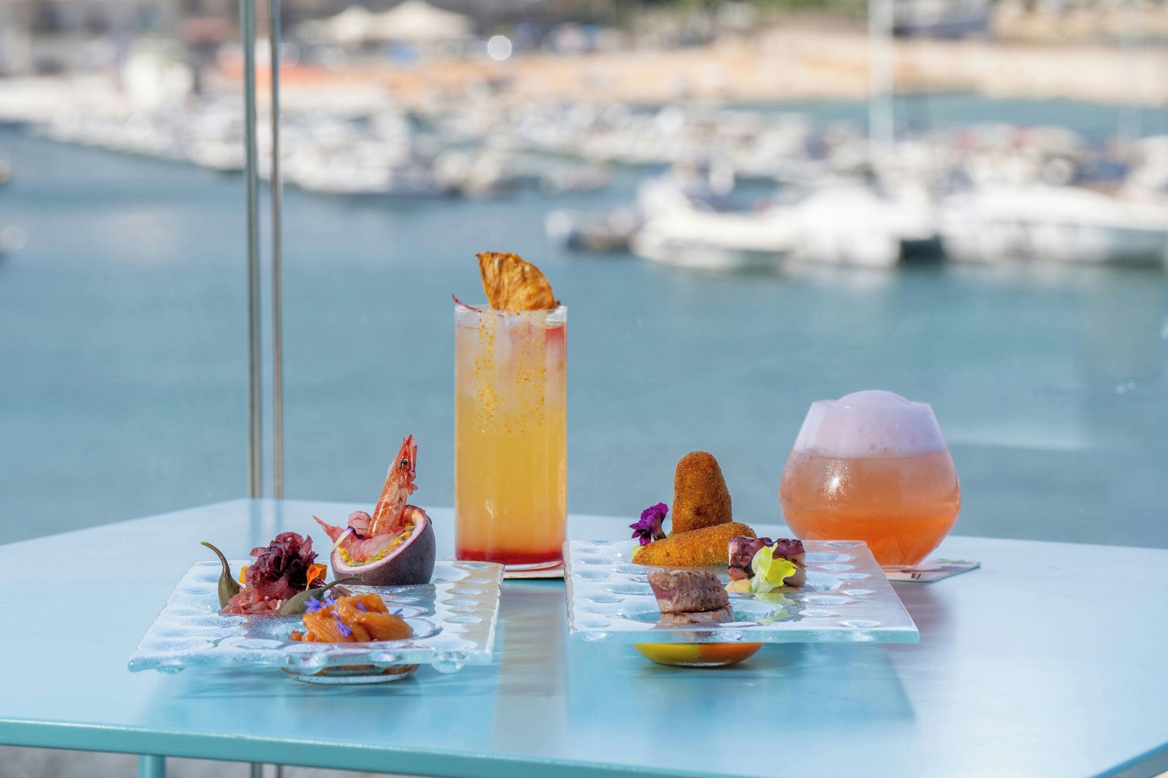 Ein Tisch mit drei Getränken und verschiedenen Vorspeisen, mit Blick auf eine Marina.