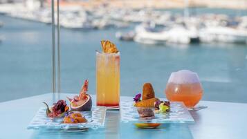 A table with three drinks and a variety of appetizers, set against a marina view.