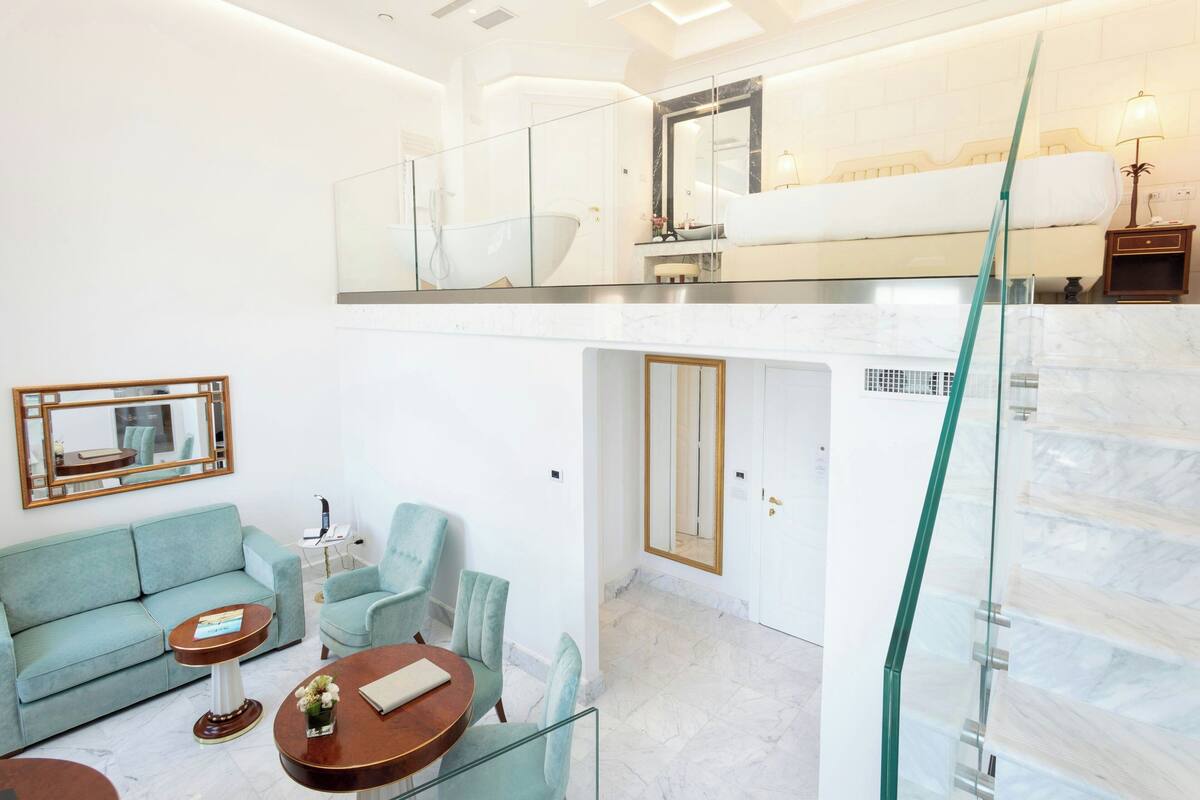 A modern hotel room with a glass staircase, a sitting area with a sofa and chairs, a small table, and a bathroom with a bathtub and a large mirror.