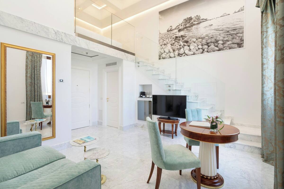 A modern living room with a marble floor, a dining area with a round table and chairs, a large flat-screen TV, and a staircase leading to an upper level.