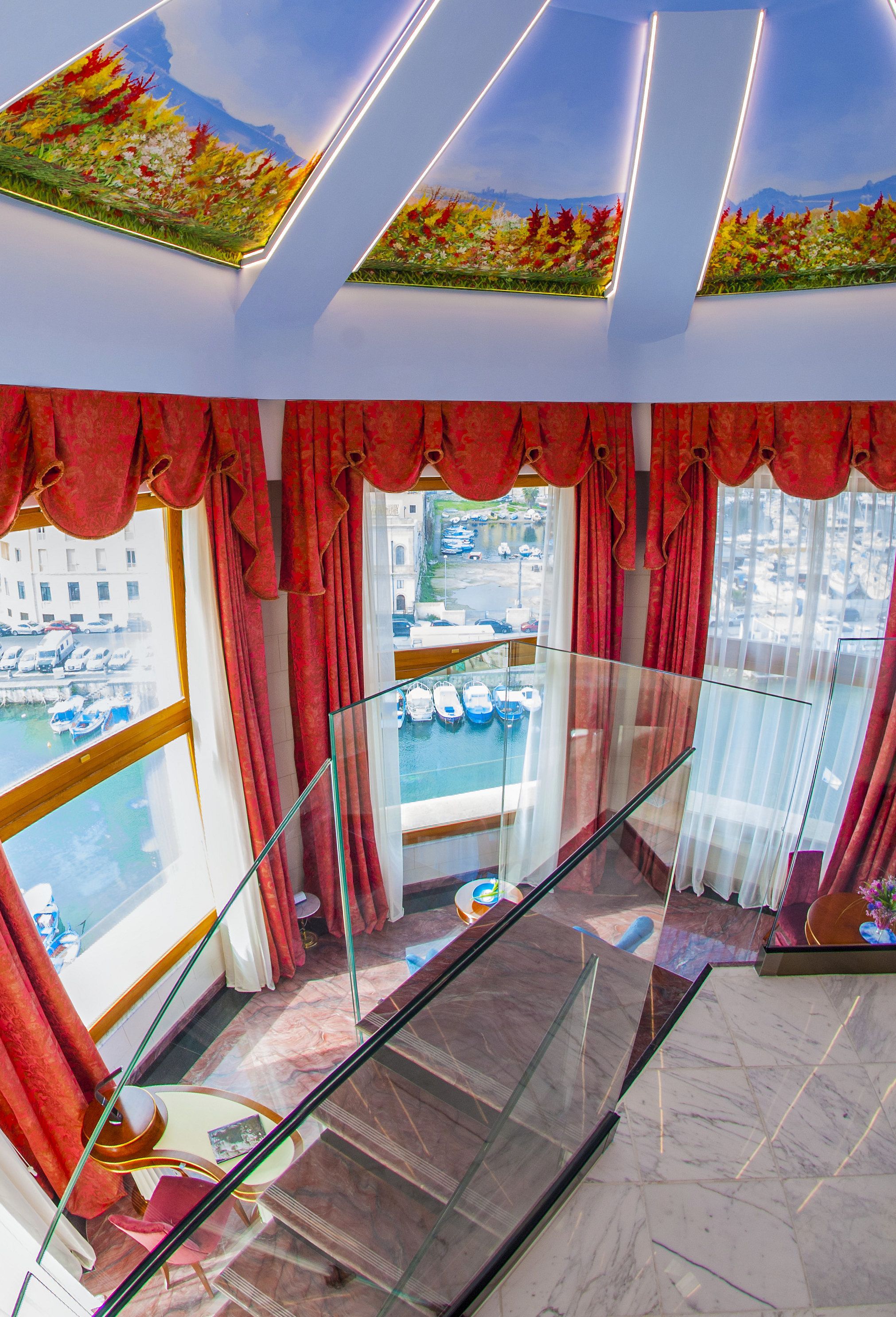 A hotel room with a balcony, red curtains, a glass railing, and a view of boats and buildings.