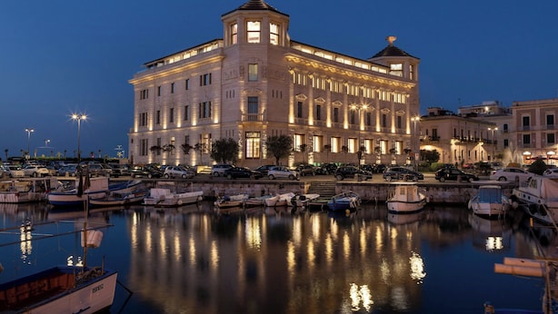 A large, historic building with multiple floors, a prominent tower, and a waterfront marina with boats. - Ortea Palace Hotel, Sicily, Autograph Collection (Syracuse)