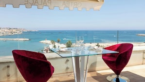 A balcony with a glass table and two red chairs, overlooking a coastal view with buildings and boats. - Ortea Palace Hotel, Sicily, Autograph Collection (Syracuse)