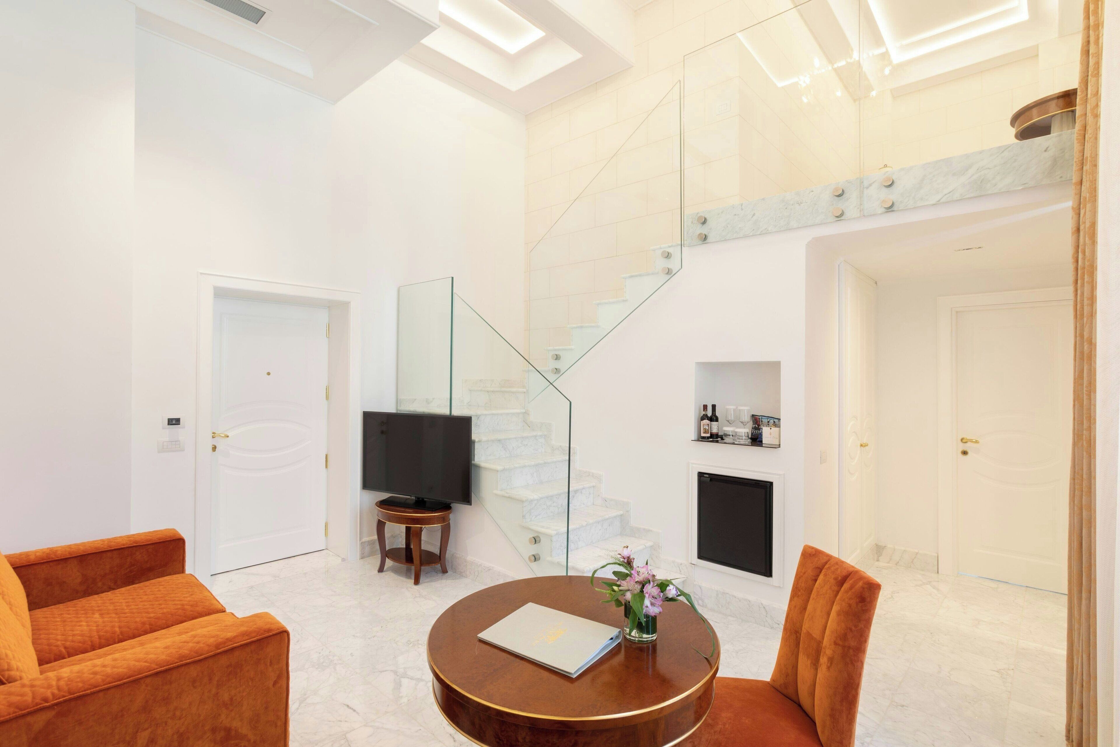 Un salon moderne avec un sol en marbre, une table basse ronde en bois, deux canapés orange, un escalier en verre et un téléviseur à écran plat.