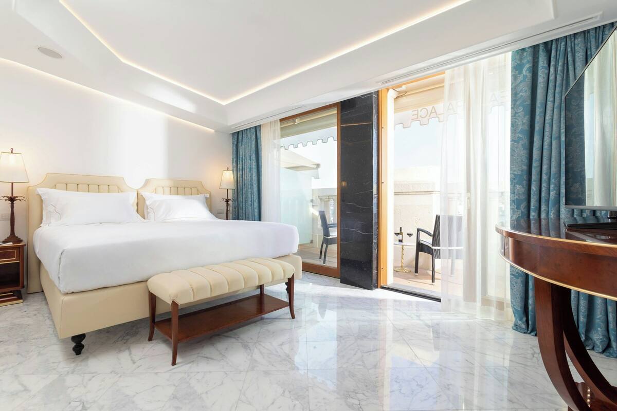 A modern hotel room with a large bed, a bench, a desk, and a view of the outdoor area.