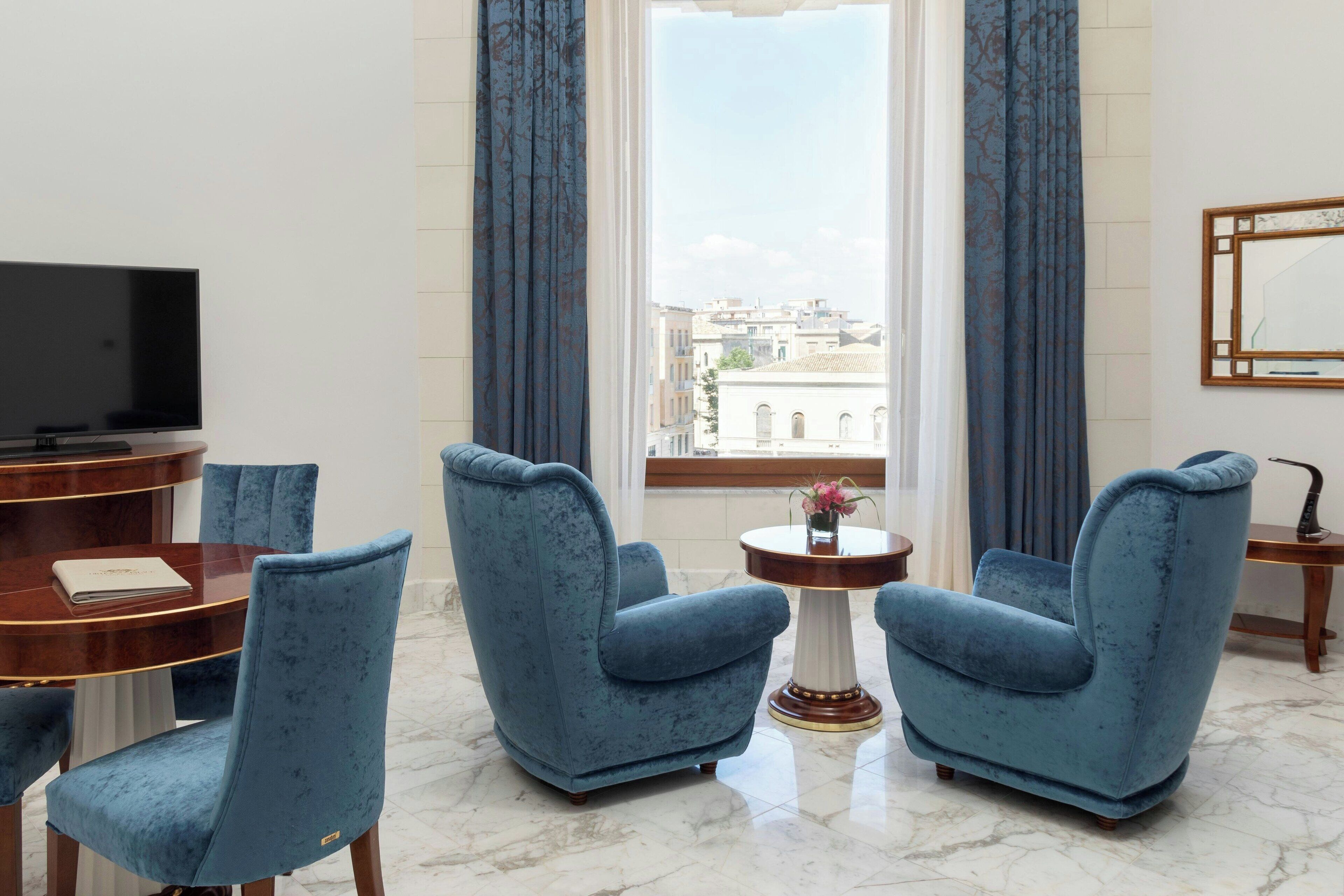 Ein modernes Wohnzimmer mit Fernseher, einem Esstisch, zwei blauen Sesseln, einem kleinen Beistelltisch mit einer Vase und Blick auf Gebäude durch ein Fenster mit blauen Vorhängen.