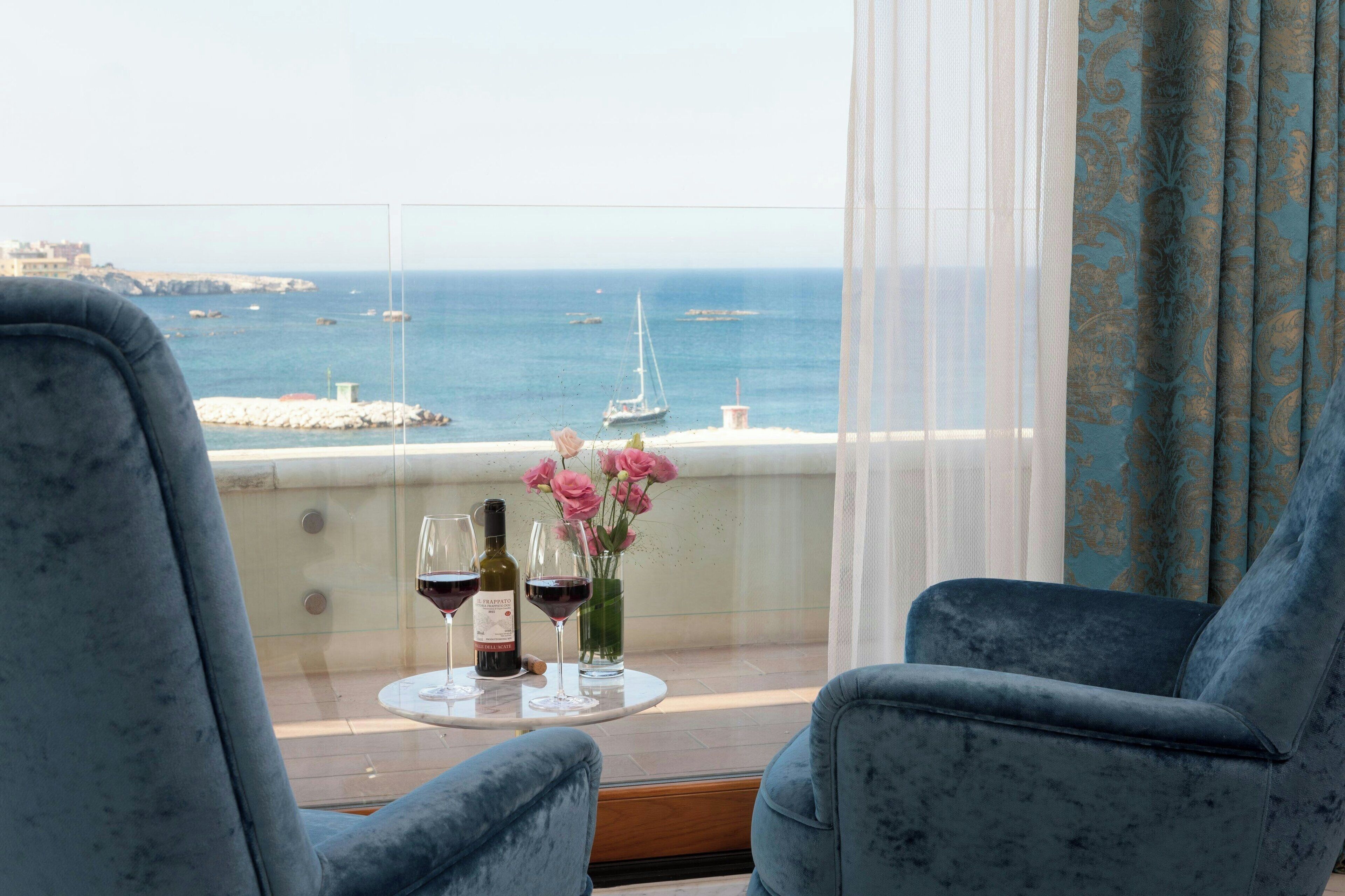 A balcony with two blue armchairs, a small table with wine glasses and flowers, and a view of the sea with boats.