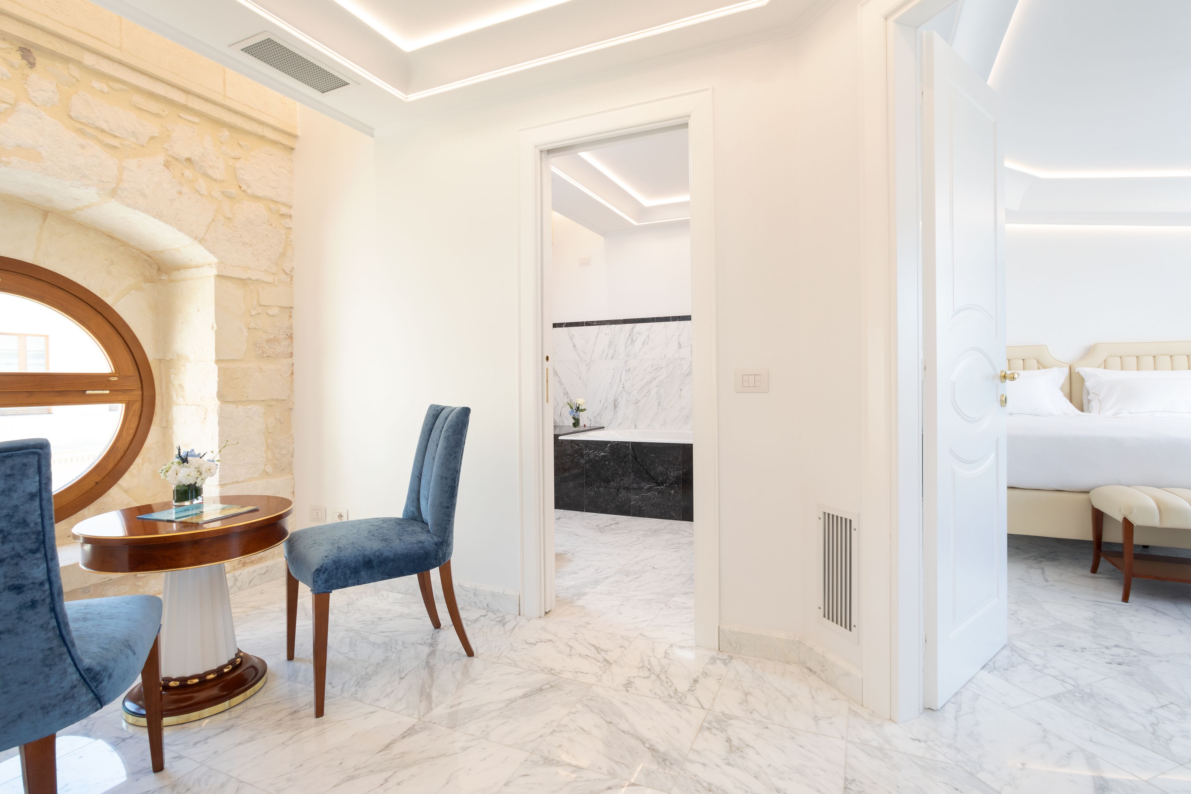 Une chambre d’hôtel moderne avec un sol en marbre, une table à manger ronde entourée de chaises bleues et un lit recouvert de draps blancs.