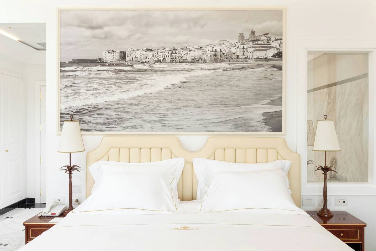 A hotel room with a bed, two bedside lamps, a nightstand with a phone, and a large black and white photograph of a coastal town on the wall.