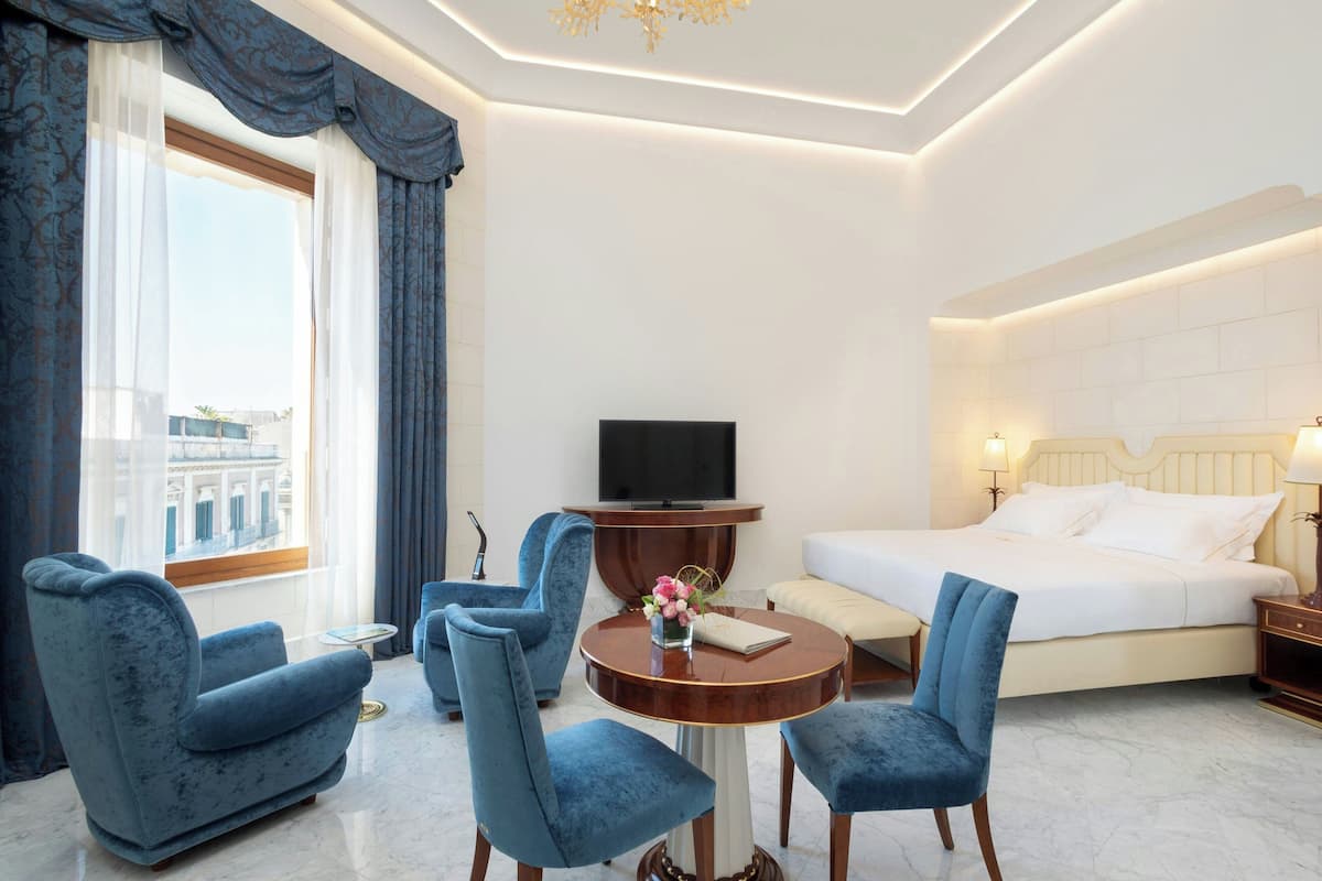 A hotel room with a large bed, a TV, a round table with chairs, and a view of a building outside.