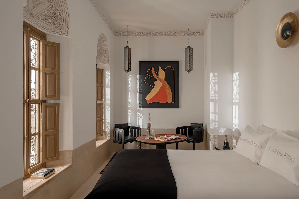 Camera d'albergo con un letto, un tavolo con bottiglia e bicchieri, due sedie e un quadro appeso al muro.