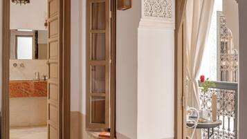 Habitación de hotel con un espejo grande, una puerta de madera, un balcón con barandilla de hierro forjado y vistas al exterior.