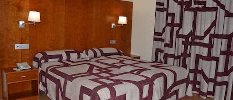 Habitación de hotel con cabecera de madera, dos lámparas de noche, colcha estampada, mesita de noche con teléfono y cortina de diseño geométrico.