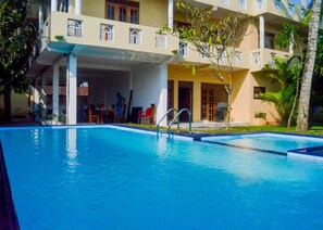A swimming pool with a clear blue water surface, surrounded by a covered outdoor area with a building in the background. - Sholay Villa (Galle)