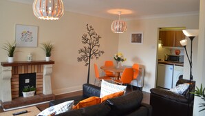 Una sala de estar con chimenea, un comedor con sillas color naranja y una cocina al fondo.
