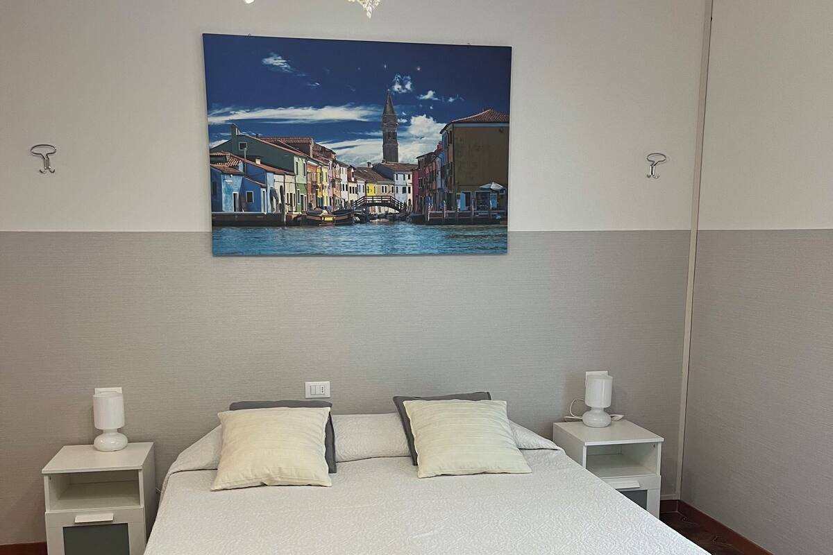 Une chambre d’hôtel avec un lit, deux tables de chevet blanches, une lampe blanche et un tableau représentant un paysage urbain.
